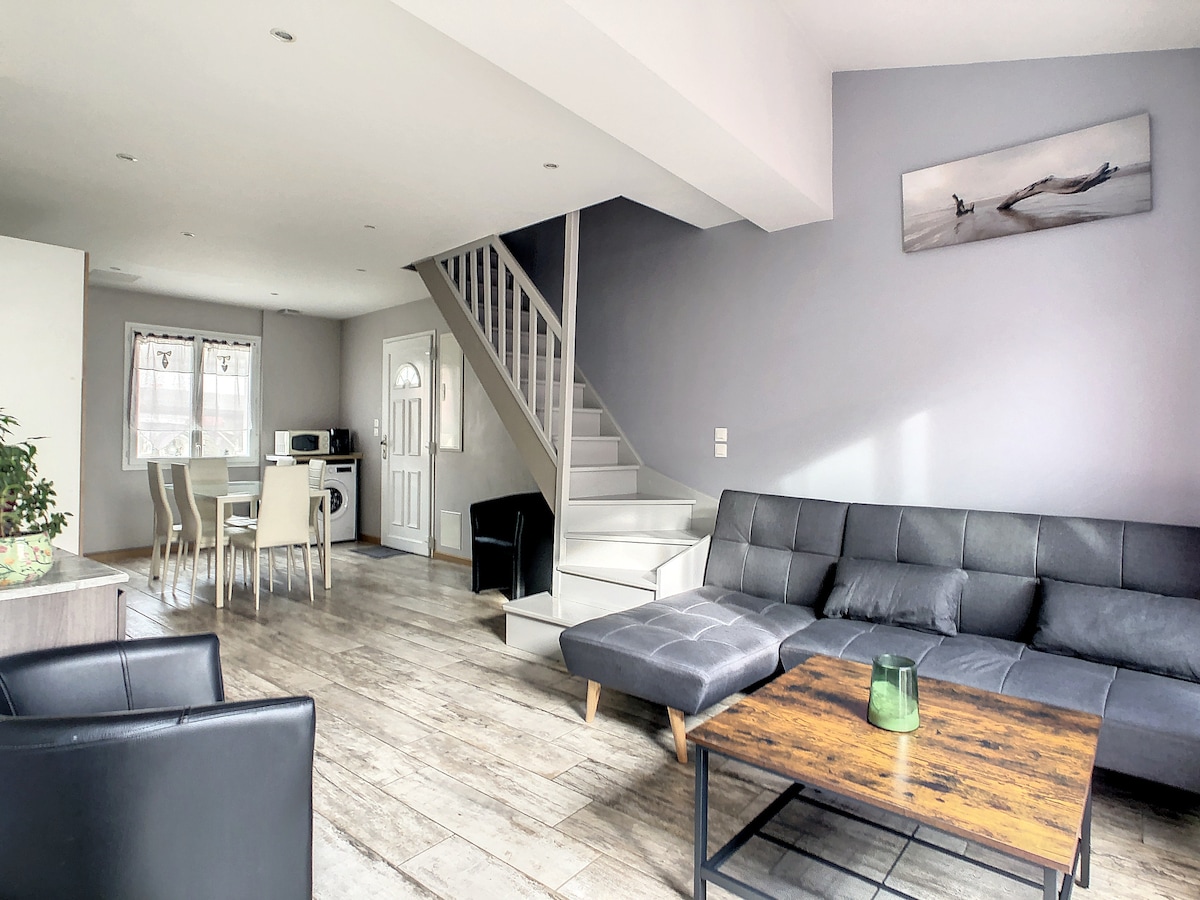 The living area features a modern layout with a sleek black sofa and a wooden coffee table at the center. A bright space is enhanced by natural light coming through the windows. A dining set is visible, along with stairs leading to the upper level.