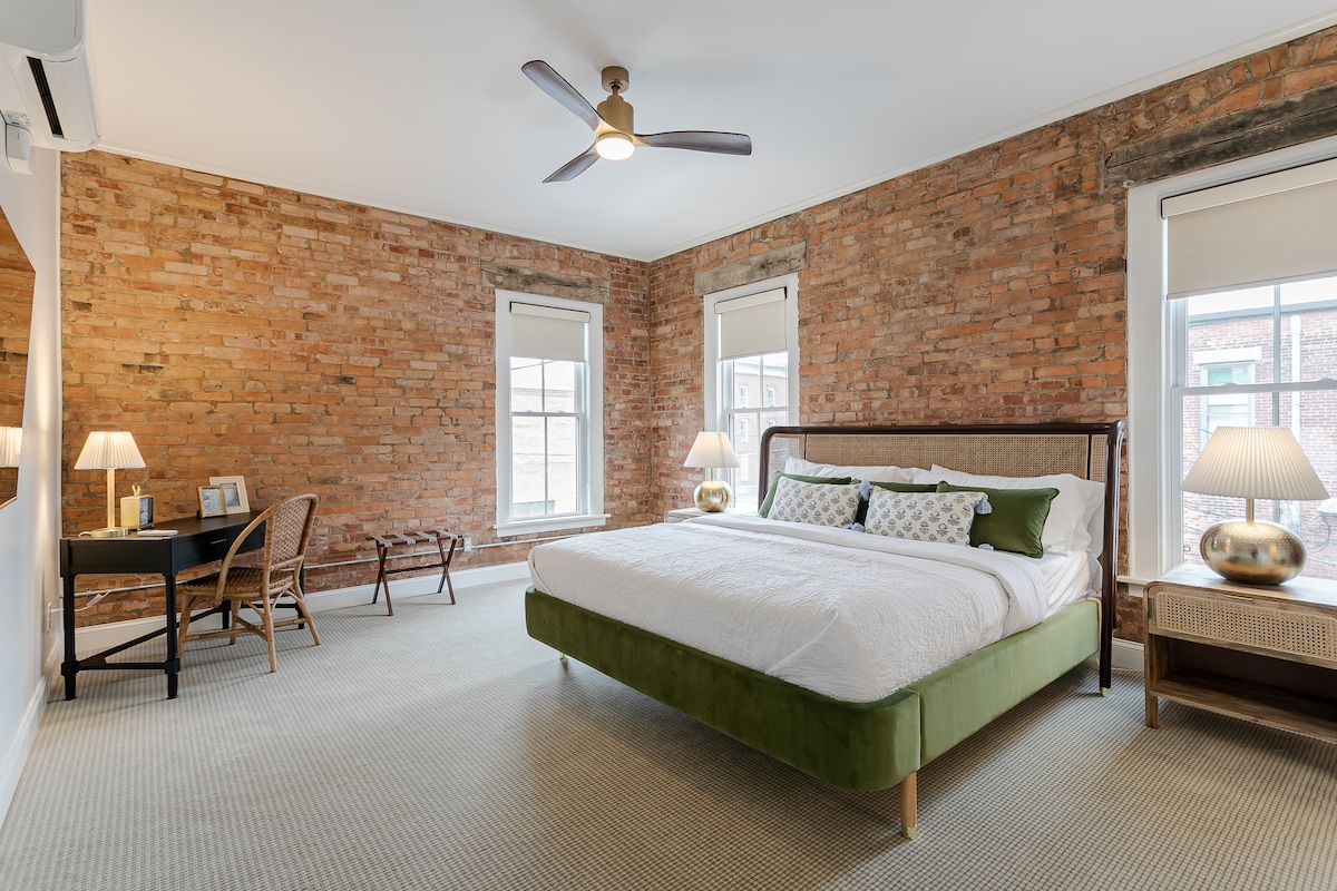 A spacious bedroom features exposed brick walls and large windows that invite natural light. A comfortable bed with decorative pillows is complemented by a stylish desk and seating area. Soft lighting from bedside lamps enhances the inviting environment.