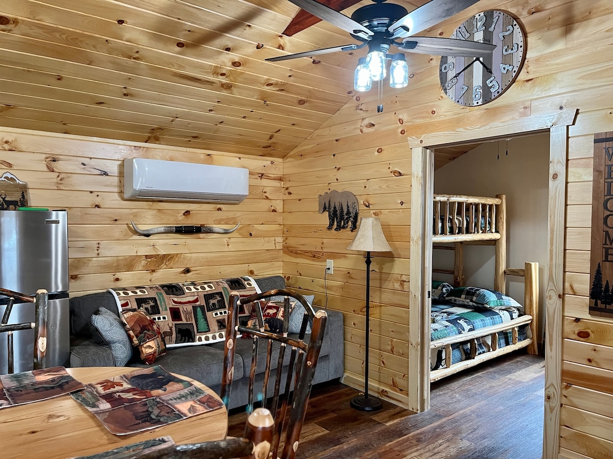 An inviting cabin interior features natural wood paneling and a ceiling fan. A cozy couch is positioned adjacent to a rustic dining table. Bunk beds are visible in a separate nook, adorned with plaid bedding, contributing to a warm and welcoming atmosphere.