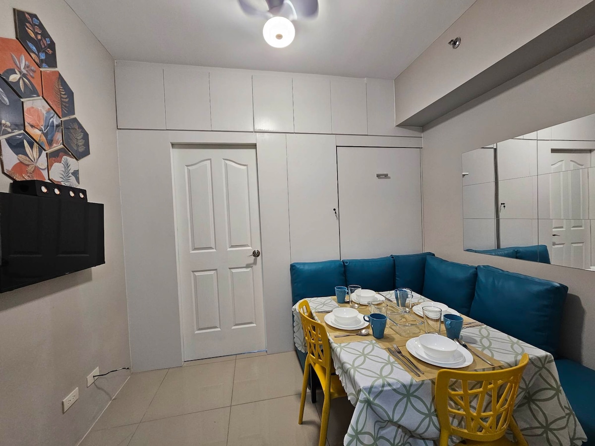 A dining area features a table set for four, accompanied by yellow chairs and a light-colored tablecloth. A blue upholstered seating area is positioned against the wall. A mirror reflects the space, while artwork is displayed on the adjacent wall, enhancing the room's design.