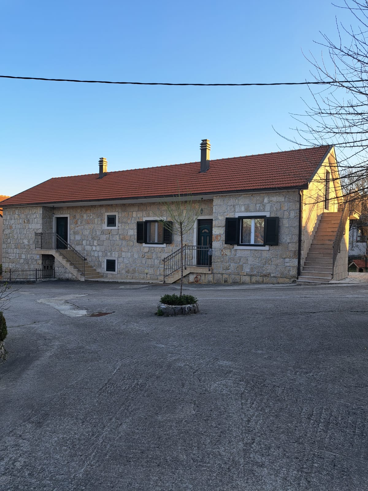 A two-story house features a traditional stone façade with a sloped roof. Two sets of stairs lead to the main entrance, which is framed by dark shutters. The surrounding area is paved, offering easy access for guests.