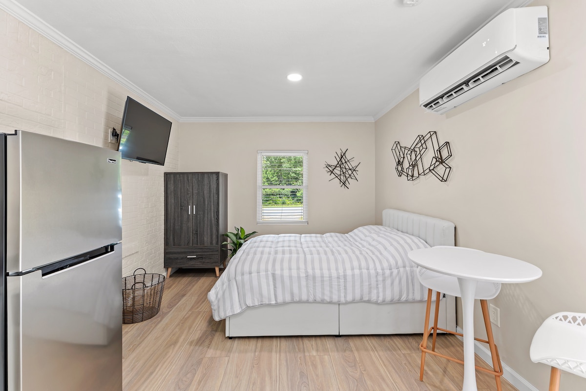 A well-appointed studio apartment features a spacious bed dressed in striped linens. A small white table and wooden chair provide a dining option. A modern television is mounted on the wall, with a compact refrigerator and dark wooden storage unit visible in the room.