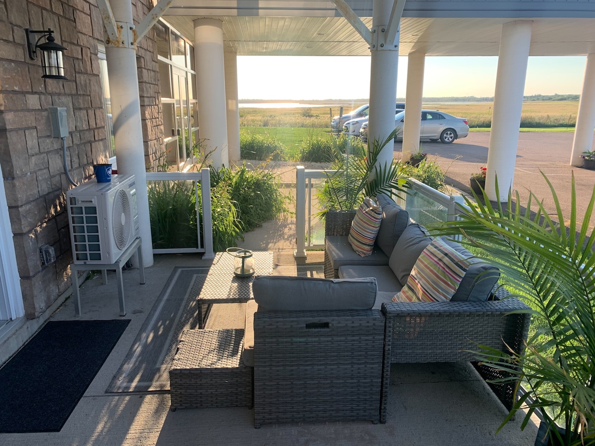 A covered outdoor seating area features a grey wicker sofa adorned with colorful cushions. A small coffee table is positioned in front of the seating, surrounded by greenery and potted plants. The soft morning light illuminates the open space, offering views of the peaceful landscape beyond.