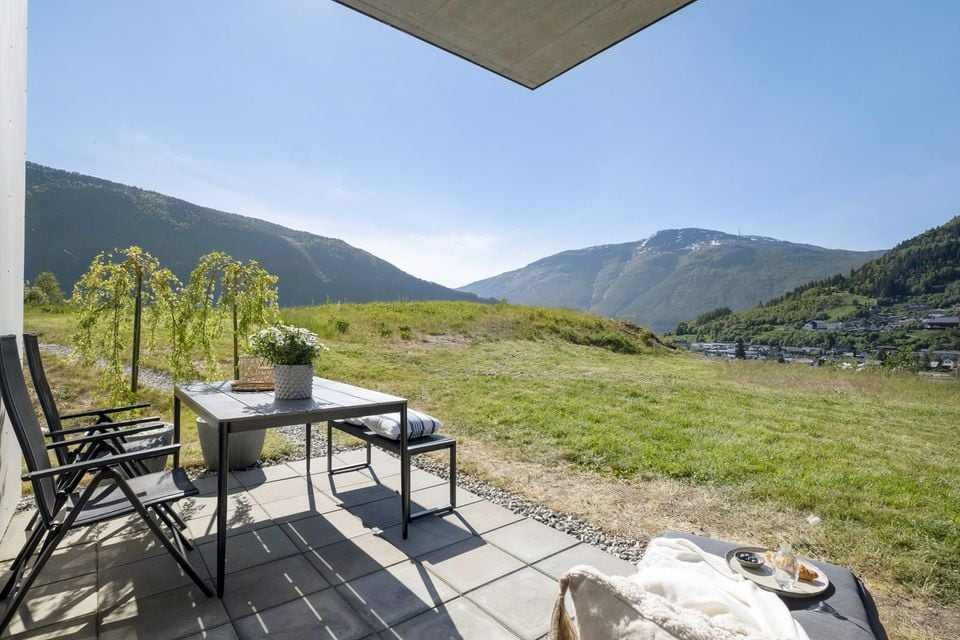 A spacious outdoor area is presented with a table and chairs arranged on a stone-paved patio. The surrounding landscape features rolling hills under a clear blue sky, with a lush green lawn enhancing the natural setting.