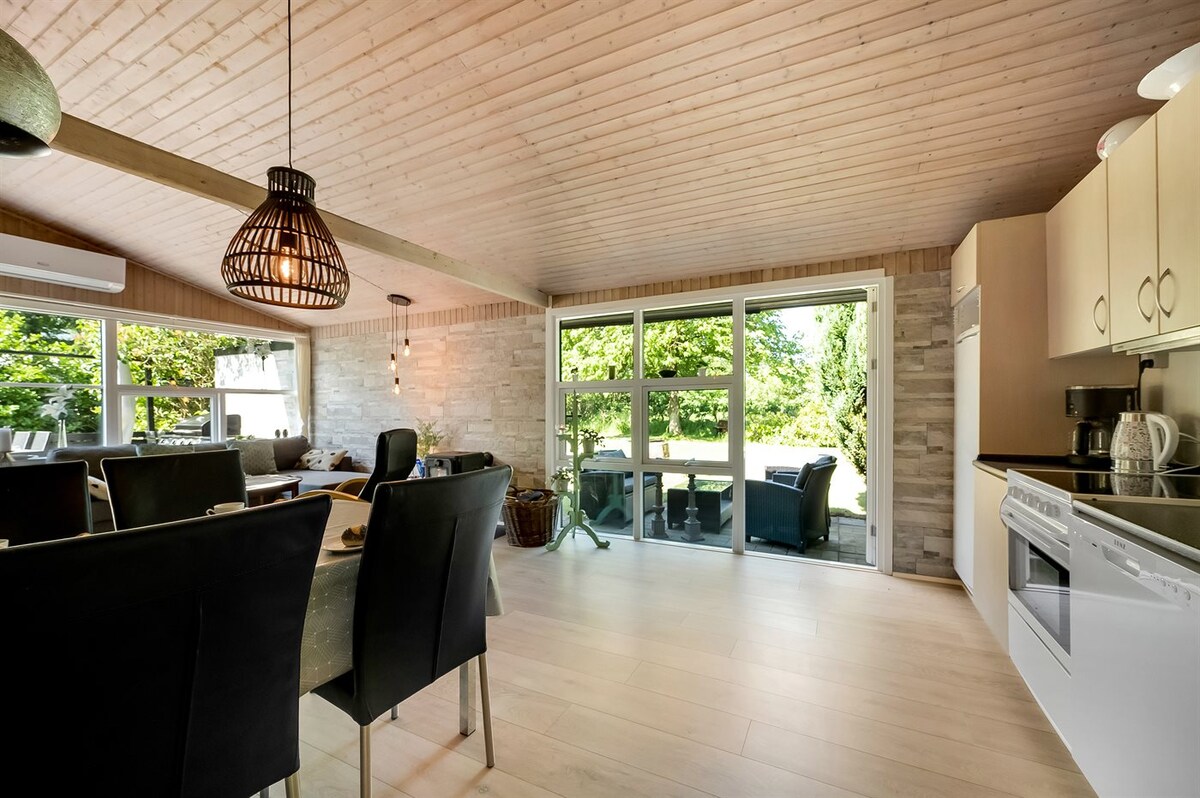 An open-plan kitchen and dining area features large glass doors that provide a view of the garden. A wooden ceiling adds warmth, while a dining table accommodates several chairs. The kitchen includes essential appliances and ample counter space, ensuring a functional living environment.