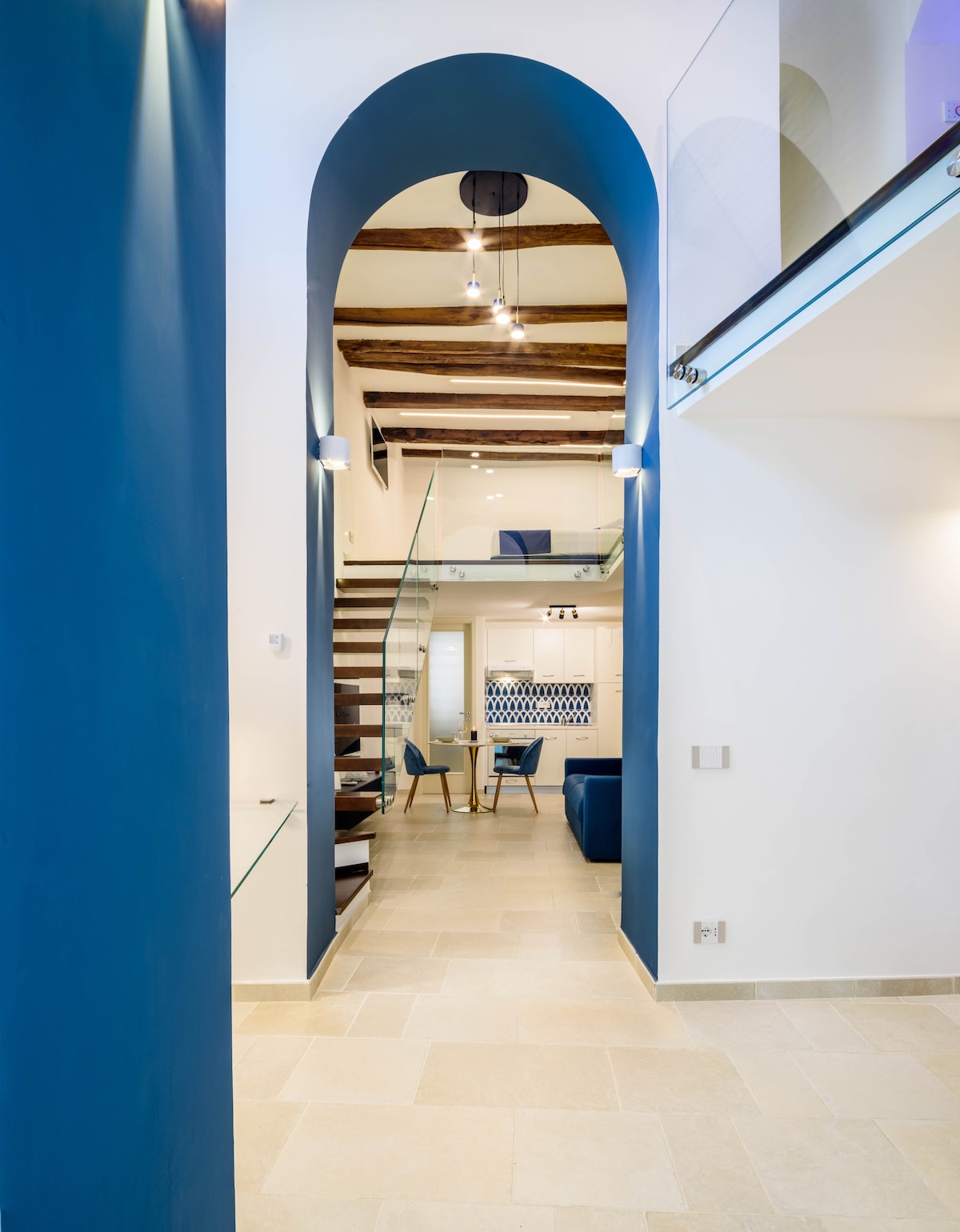 A modern entryway features a high ceiling with exposed wooden beams, creating an airy feel. A striking blue archway frames the view into the living area, where stylish furnishings are visible. Illuminated artwork hangs on the wall, enhancing the design.