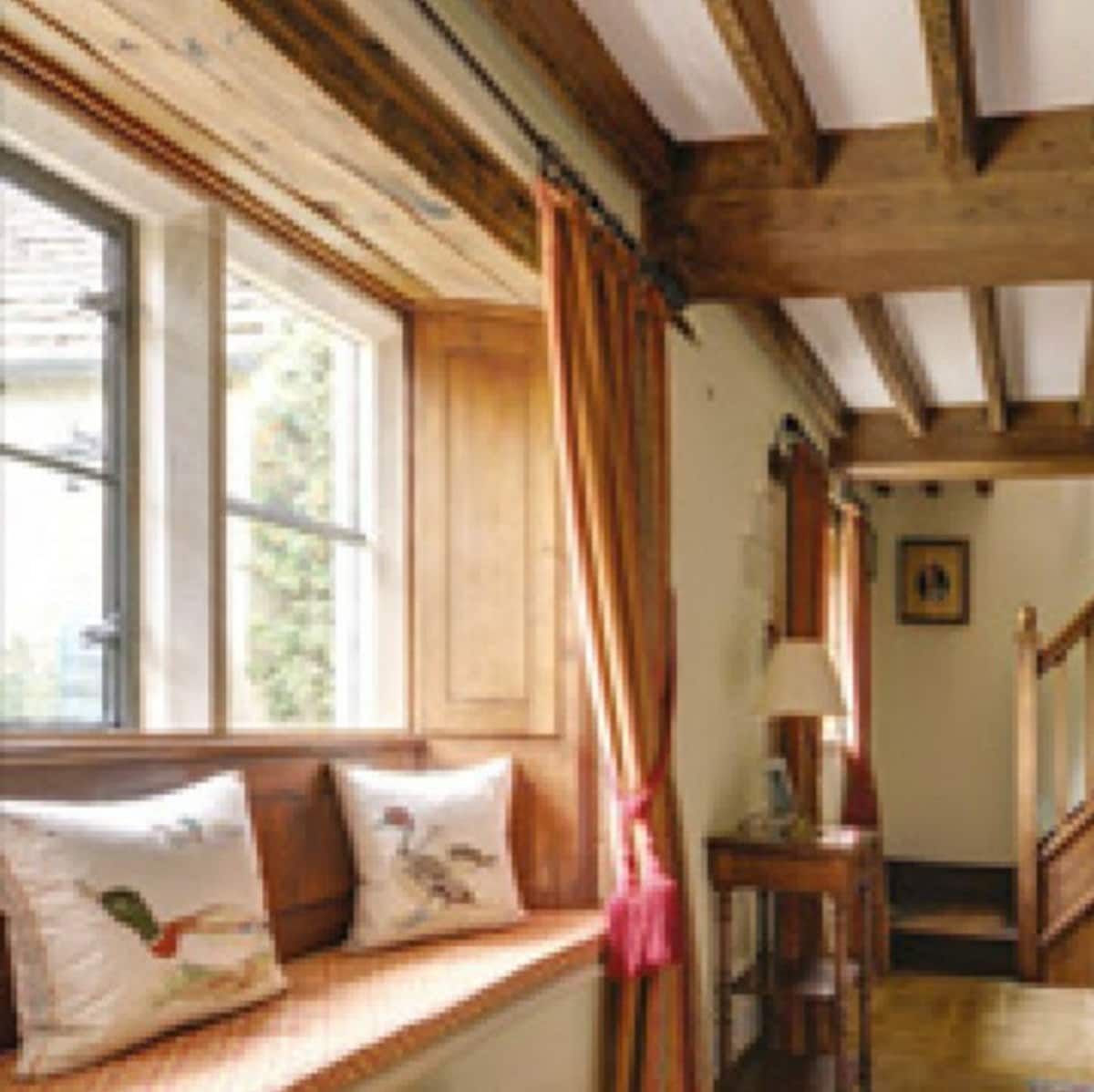 A charming hallway features wooden beams and large windows with decorative cushions on a built-in bench. Soft curtains frame the windows, allowing natural light to illuminate the space. A wooden table sits against the wall, contributing to the cottage's welcoming atmosphere.