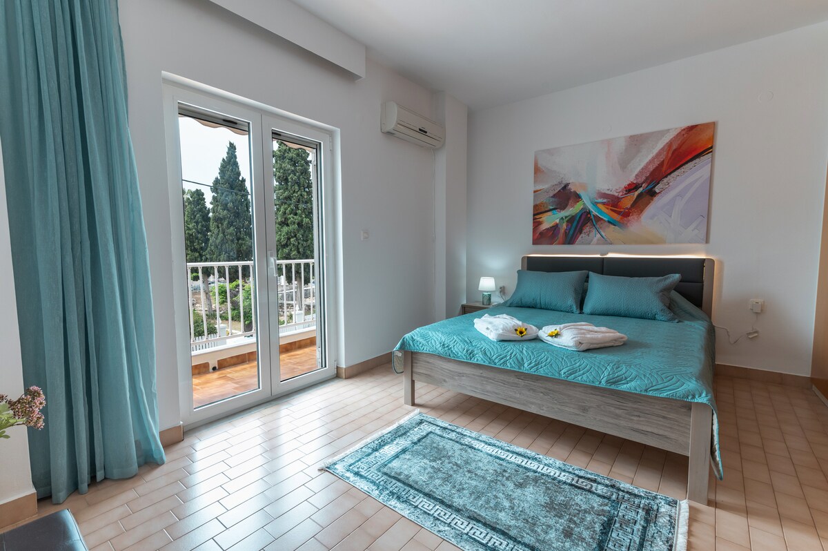 A spacious bedroom features a large bed dressed in a vibrant teal coverlet. Natural light enters through the glass balcony doors, illuminating the area. A colorful abstract artwork adorns the wall above the bed, while a soft rug lies beneath it. Air conditioning is present.