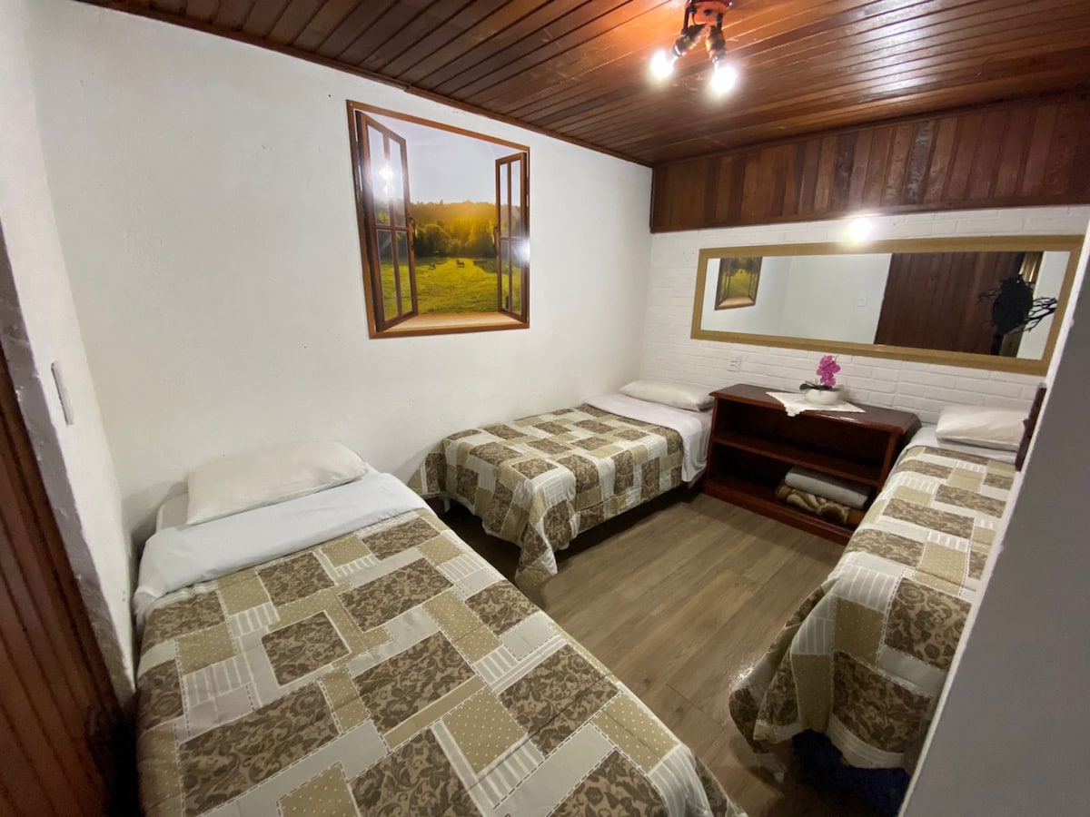 A spacious room is equipped with three beds, each adorned with patterned bedding. A wooden console is positioned against the wall, complemented by a decorative mirror. Natural light filters through a window framed by a scenic landscape photograph.
