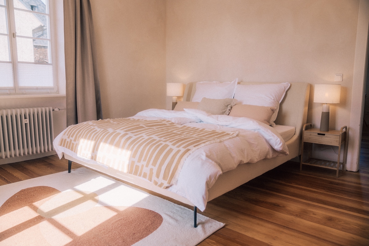 A spacious bedroom features a large bed with soft bedding in neutral tones. Light streams through a window, illuminating the wooden floor. Two bedside lamps provide subtle lighting, and a decorative rug adds a touch of warmth to the space.