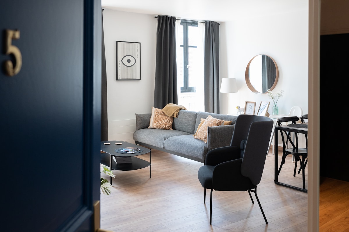 A spacious and bright living area is displayed, featuring a large gray sofa adorned with decorative cushions. A dark round coffee table sits in front, and a round mirror reflects natural light from the window beside it. A dining table with chairs is visible in the background.