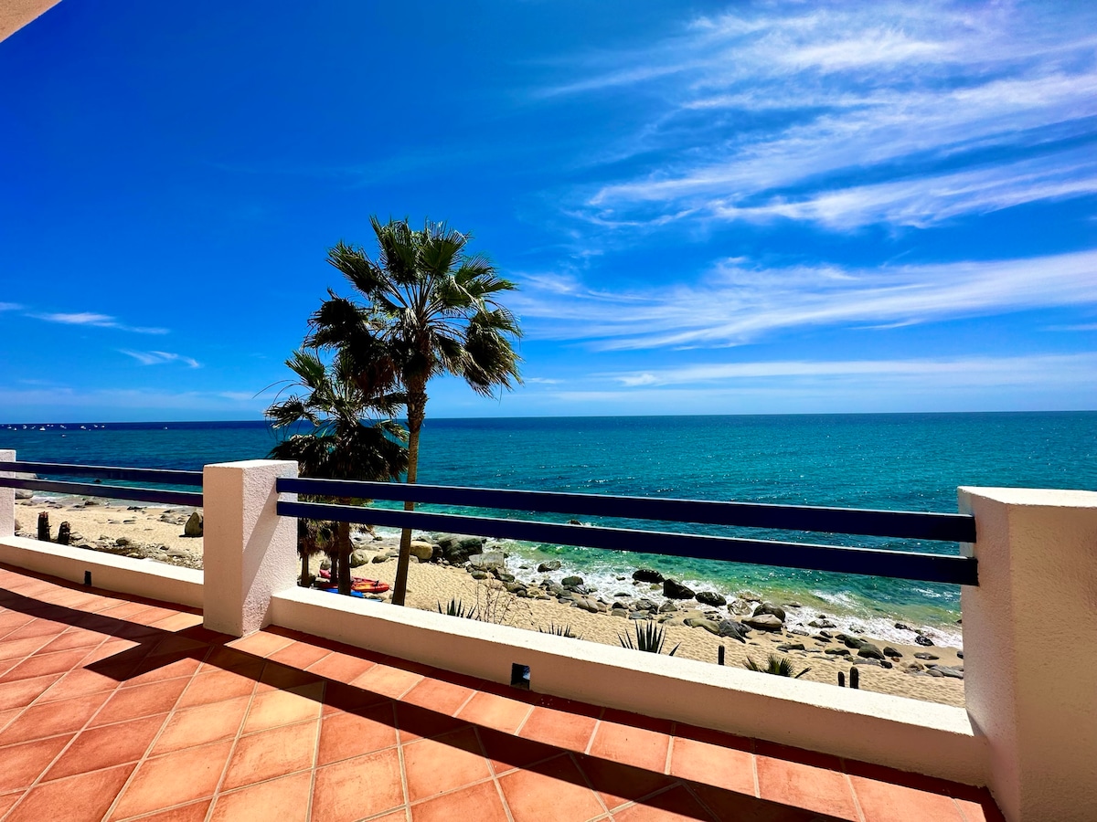 A balcony offers an expansive view of the ocean and sandy beach, framed by a palm tree. The smooth, light tile flooring contrasts with the deep blue sky and water, creating a serene coastal setting. Waves can be seen gently lapping at the shore.