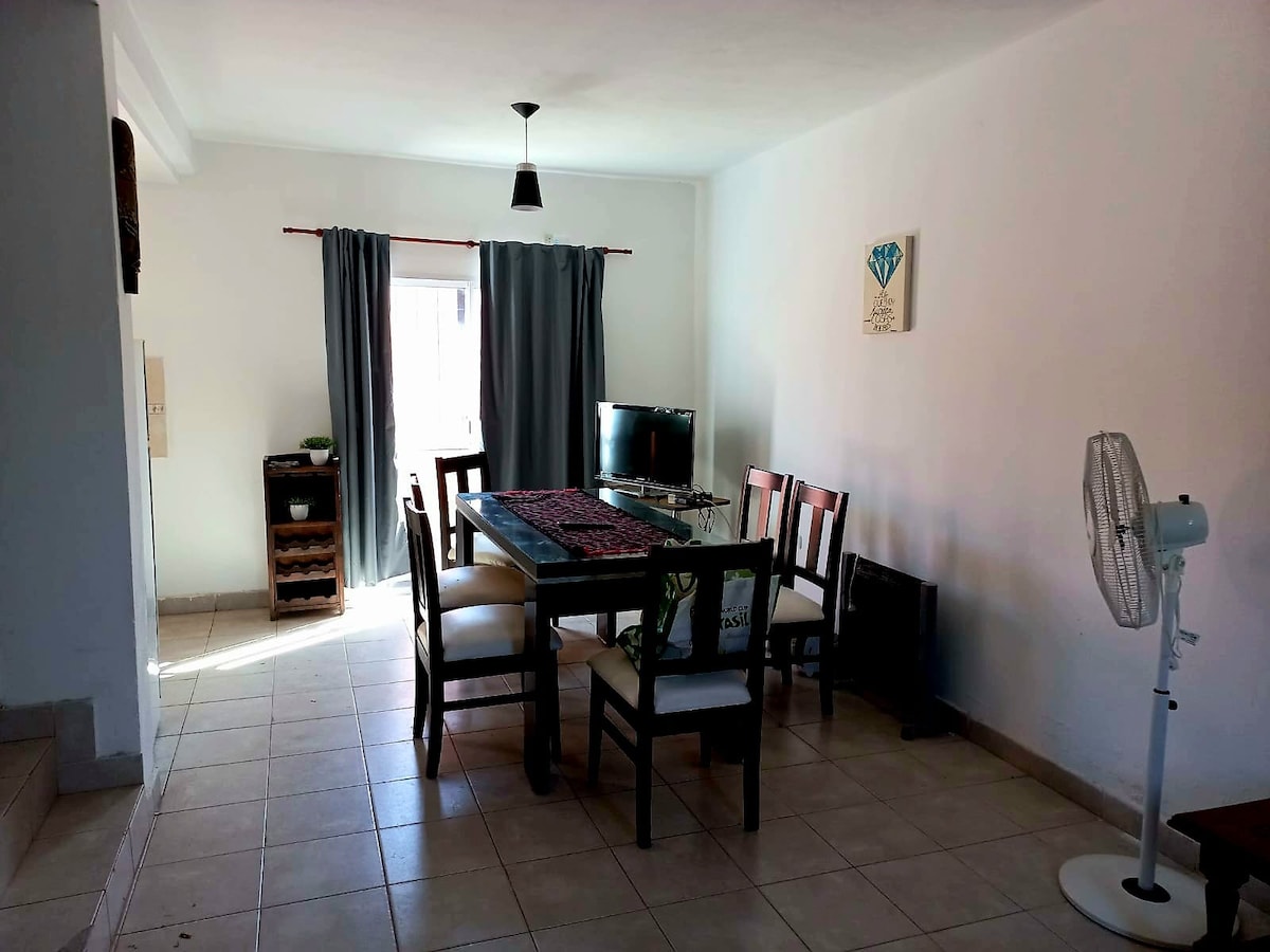 An airy dining area features a wooden table surrounded by six chairs, complemented by a patterned runner. A television is mounted on the wall, while a fan sits nearby. Natural light enters through the window, dressed with light curtains, enhancing the spacious feel.