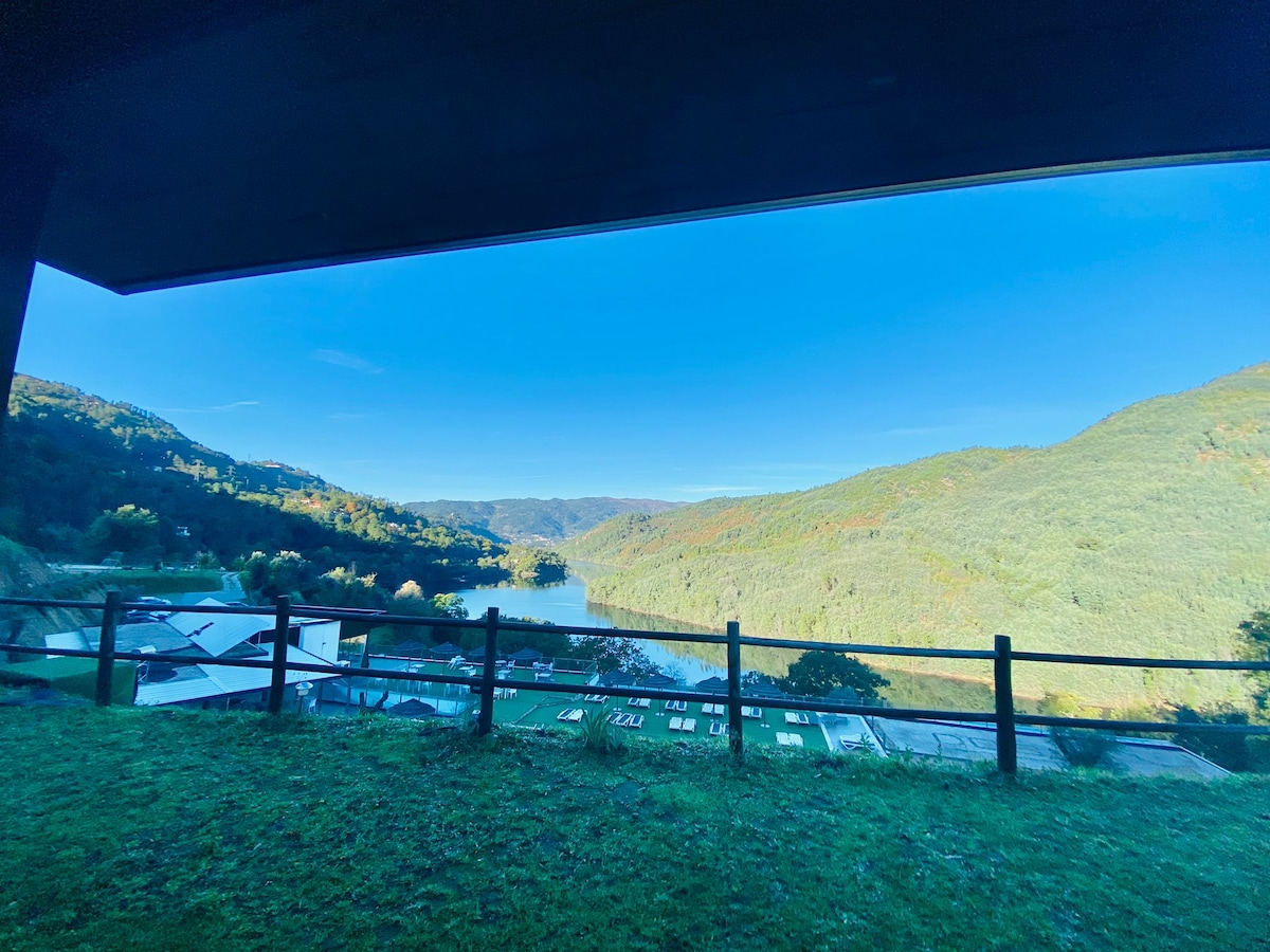 A scenic view is captured from a covered terrace, showcasing rolling hills and a calm river below. The landscape is framed by a black railing and features lush greenery under a clear blue sky.