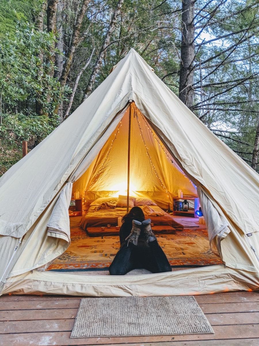 An inviting canvas bell tent is positioned on a wooden platform surrounded by trees. Inside, two twin beds with a shared nightstand and warm lighting create a comfortable sleeping area. A natural mat is placed at the tent entrance, welcoming guests.