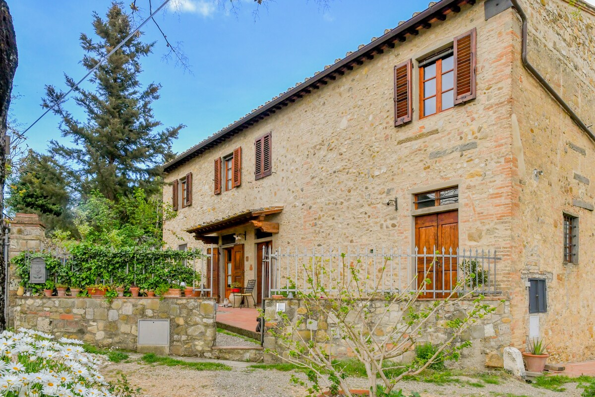 A rustic stone house is set against a backdrop of trees and greenery. The facade features traditional windows with wooden shutters, and a welcoming entrance is adorned with potted plants. The surrounding area includes a garden, enhancing the tranquil country setting.