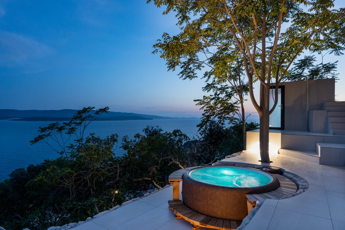 A tranquil outdoor space features a round hot tub, set on a wooden deck, surrounded by lush greenery. The scene overlooks the calm waters of the Adriatic coast during twilight, with soft lighting highlighting the natural beauty of the area.
