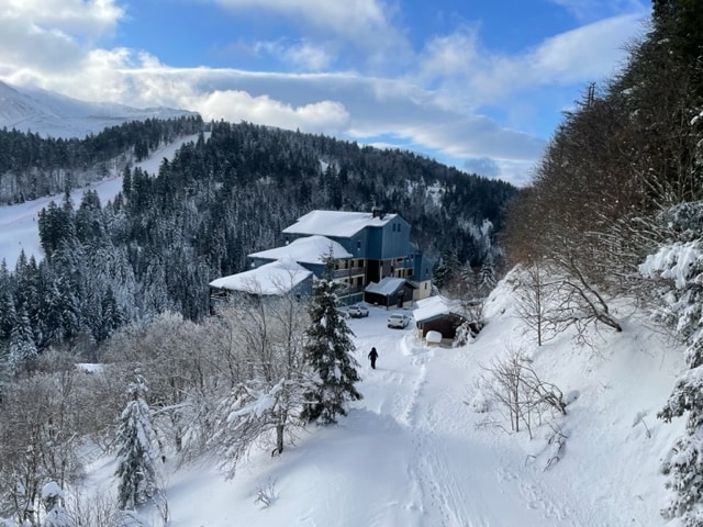 The snow-covered landscape highlights a blue building nestled among trees, with expansive mountains in the background. A person is seen walking along a snowy path, creating a serene winter scene, while the sky is filled with clouds and patches of blue.