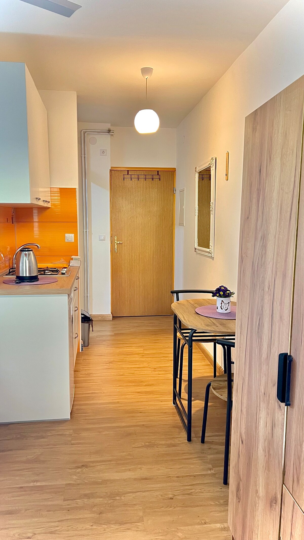 A compact kitchen area is visible, featuring modern appliances and an orange backsplash. A dining space with two black chairs and a small table is situated beside the partially open wooden cabinet. A door leading to another room can be seen at the end of the hallway.