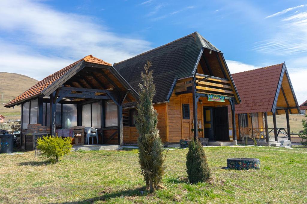 A charming wooden cabin is set against a clear sky, showcasing a sloping roof and a welcoming porch. Surrounding greenery is complemented by small shrubs and decorative stone arrangements. A secondary structure is visible, enhancing the outdoor space with a cozy, inviting vibe.