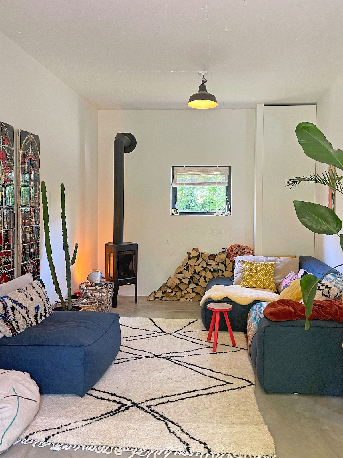 A cozy living room is presented, featuring two sofas adorned with textured cushions. A small red stool stands nearby, and a wood-burning stove is visible next to a neatly stacked pile of firewood. Natural light filters in through the window, complemented by indoor plants.