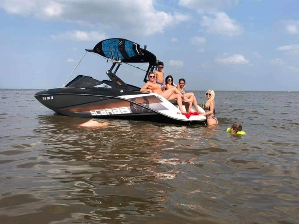 A boat is anchored in the water, with several individuals enjoying the warm day. Some are seated on the boat, while others are partially submerged in the water. The sky is slightly cloudy, reflecting a relaxed atmosphere.