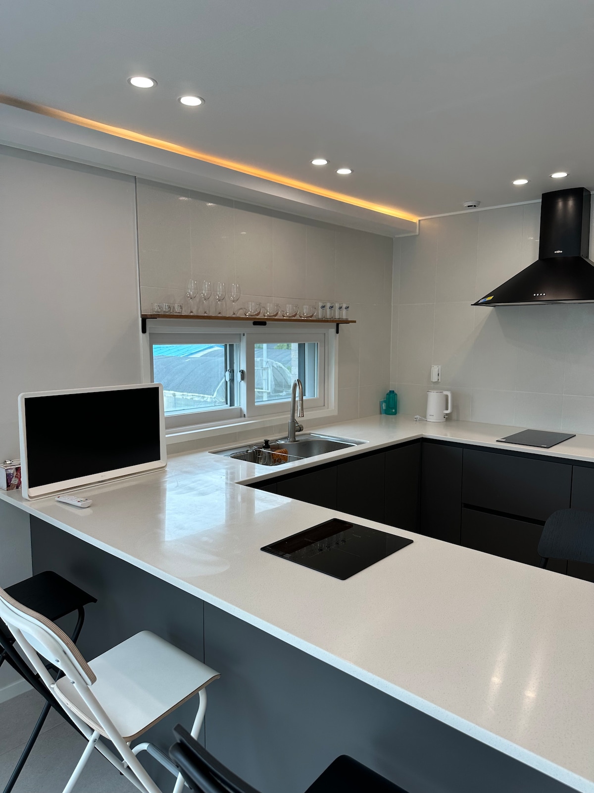 A modern kitchen is designed with an island featuring a smooth white countertop. A sleek black hood fan hangs above a stovetop. The space is illuminated by under-cabinet lighting, and a large window allows natural light to enter, complemented by glassware displayed on a shelf.