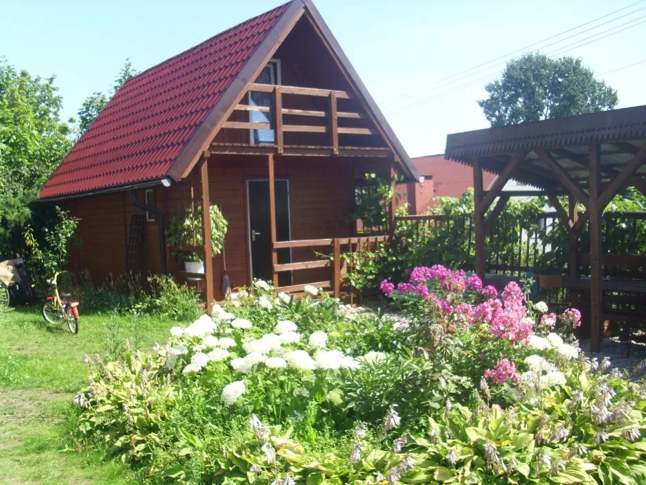 A charming wooden cottage is surrounded by a vibrant garden featuring blooming flowers and green foliage. A covered seating area is visible to the right, and a bicycle is parked nearby, providing a sense of accessibility to the outdoor space.