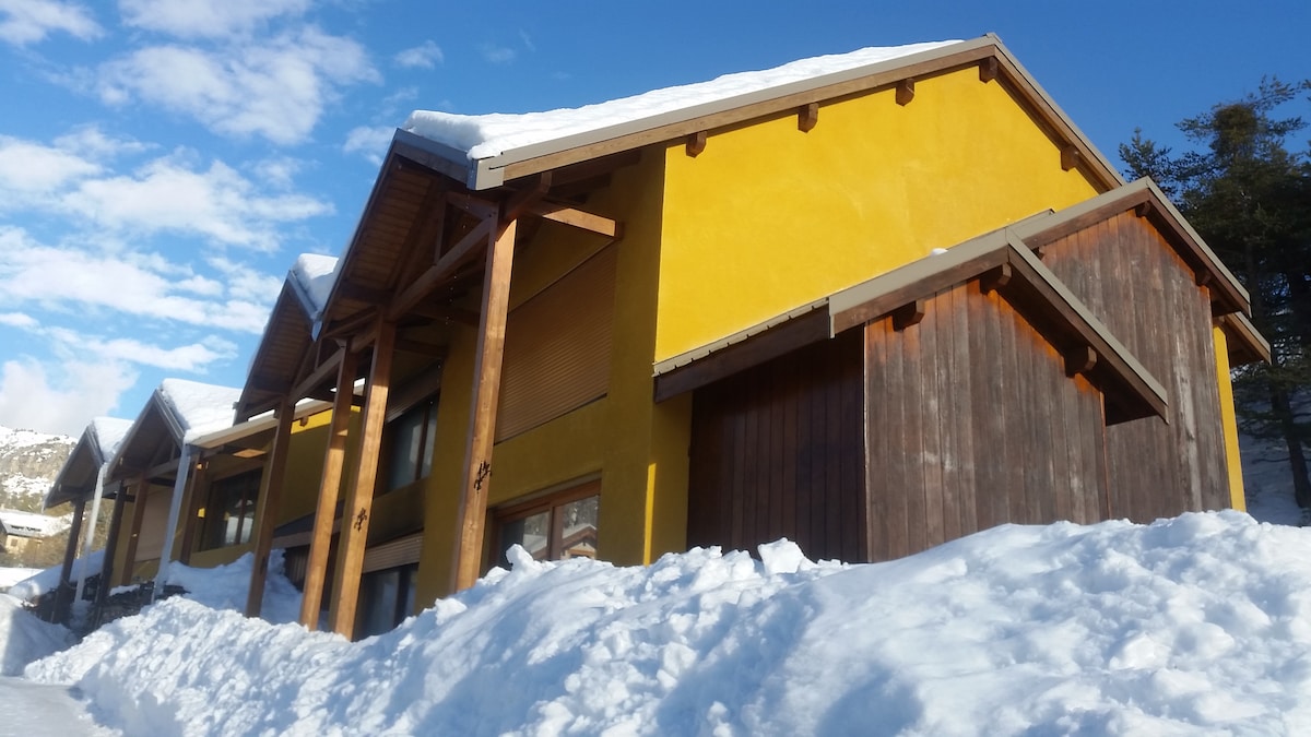 The exterior of the chalet is showcased under a clear blue sky, with yellow walls contrasting against the snow-covered ground. Wooden beams and a slanted roof add architectural interest, while the surrounding snow creates a serene winter landscape.