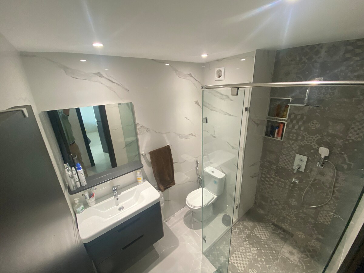 A modern bathroom features sleek cabinetry and a large mirror above a spacious sink. The shower area is enclosed with glass, showcasing contemporary tiles and a rain shower fixture. Natural light is provided by overhead recessed lighting, enhancing the clean, minimalist design.