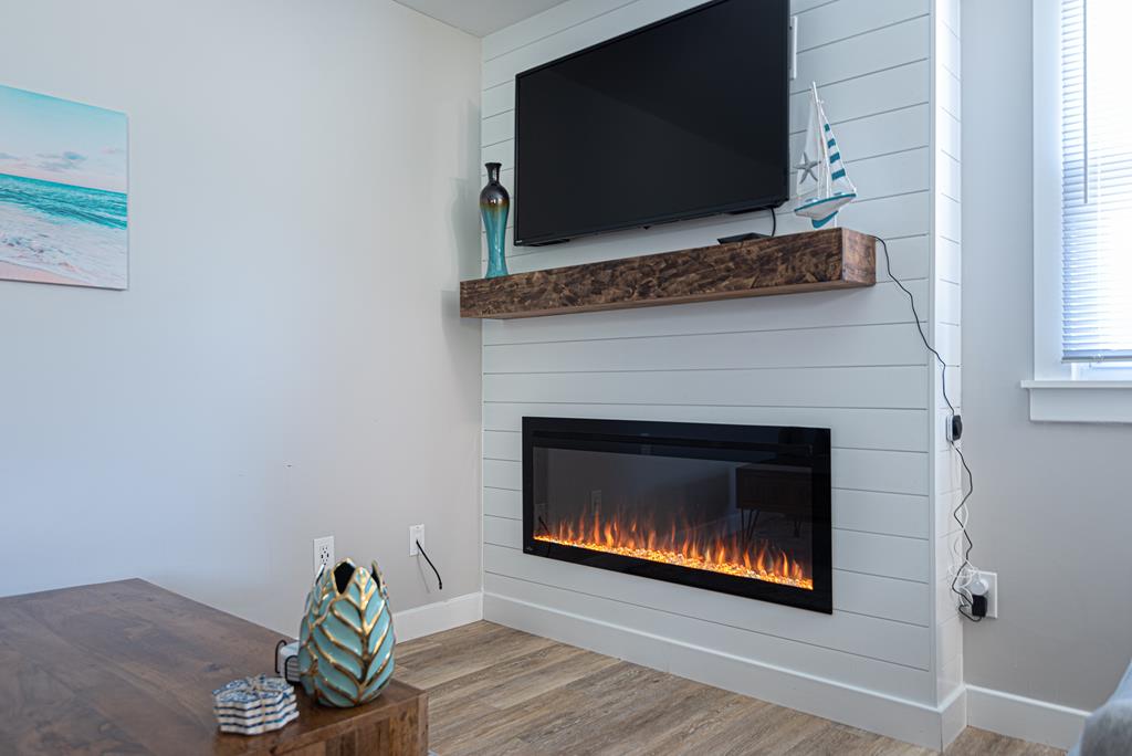 A modern living area features a wall-mounted television above a sleek wooden mantle. An electric fireplace with realistic flames adds an inviting touch. A decorative vase and a small sculpture rest on the coffee table, enhancing the space's contemporary design.