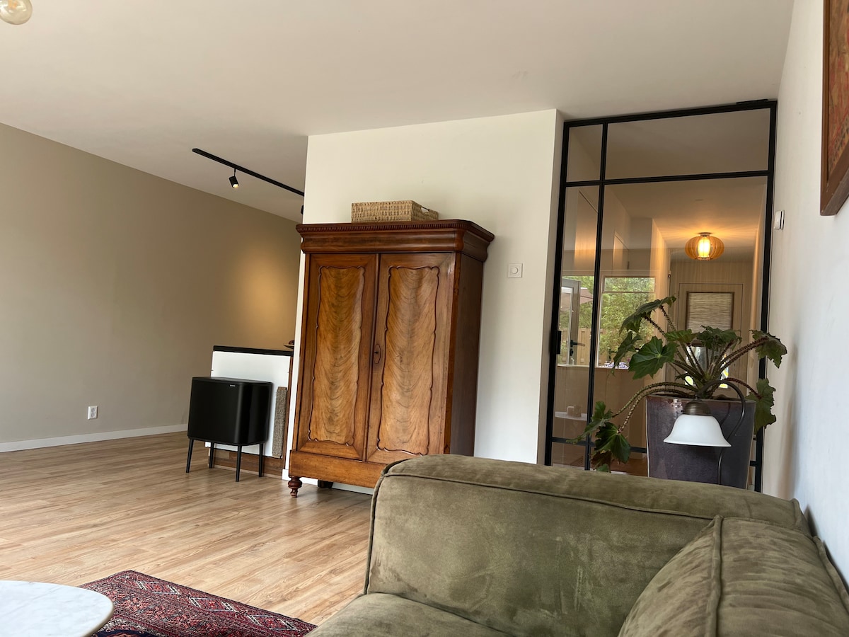 A spacious living area is highlighted by light wood flooring and neutral walls. A vintage wooden wardrobe stands against one wall, while a green couch and a round white table create a relaxed seating arrangement. Natural light enters through a nearby glass door.