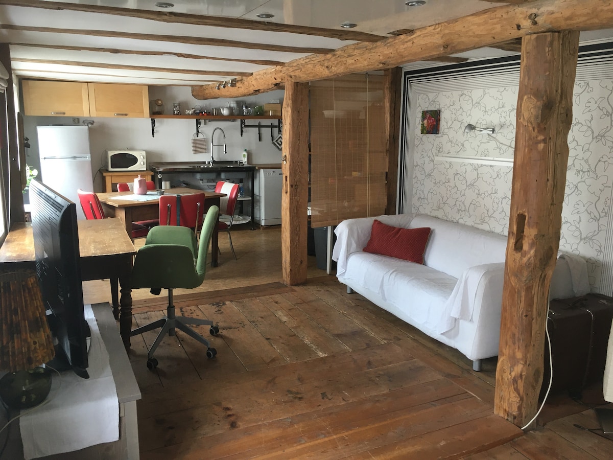 An open living space features exposed wooden beams and a wooden floor. A white sofa with a red pillow is positioned beside a green office chair and a desk. A dining table with four red chairs is visible, along with a well-equipped kitchen in the background.
