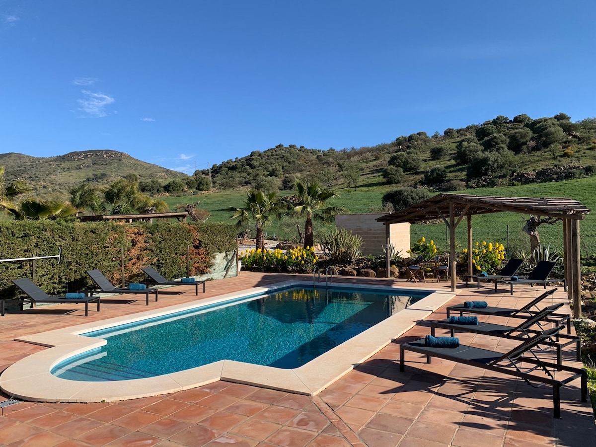 A spacious pool area is featured, surrounded by sun loungers and shaded seating under a rustic pergola. Lush greenery and vibrant yellow flowers create a natural backdrop, with hills gently rising in the distance under a clear blue sky.