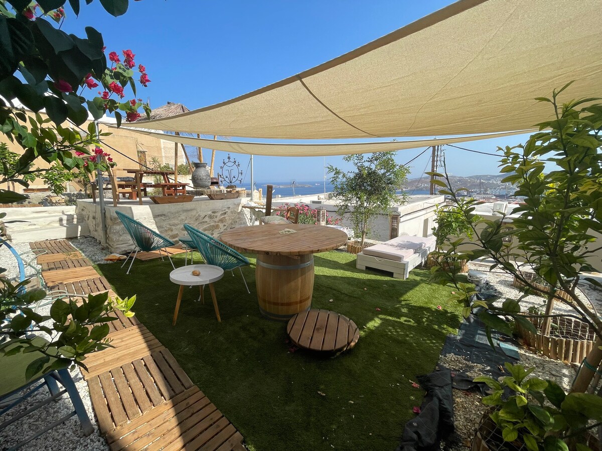An outdoor terrace is featured, with seating arrangements that include circular wooden table and green chairs. A shaded area provided by a large canopy offers a comfortable space. Lush greenery and decorative plants surround the area, with a glimpse of the sea visible in the background.