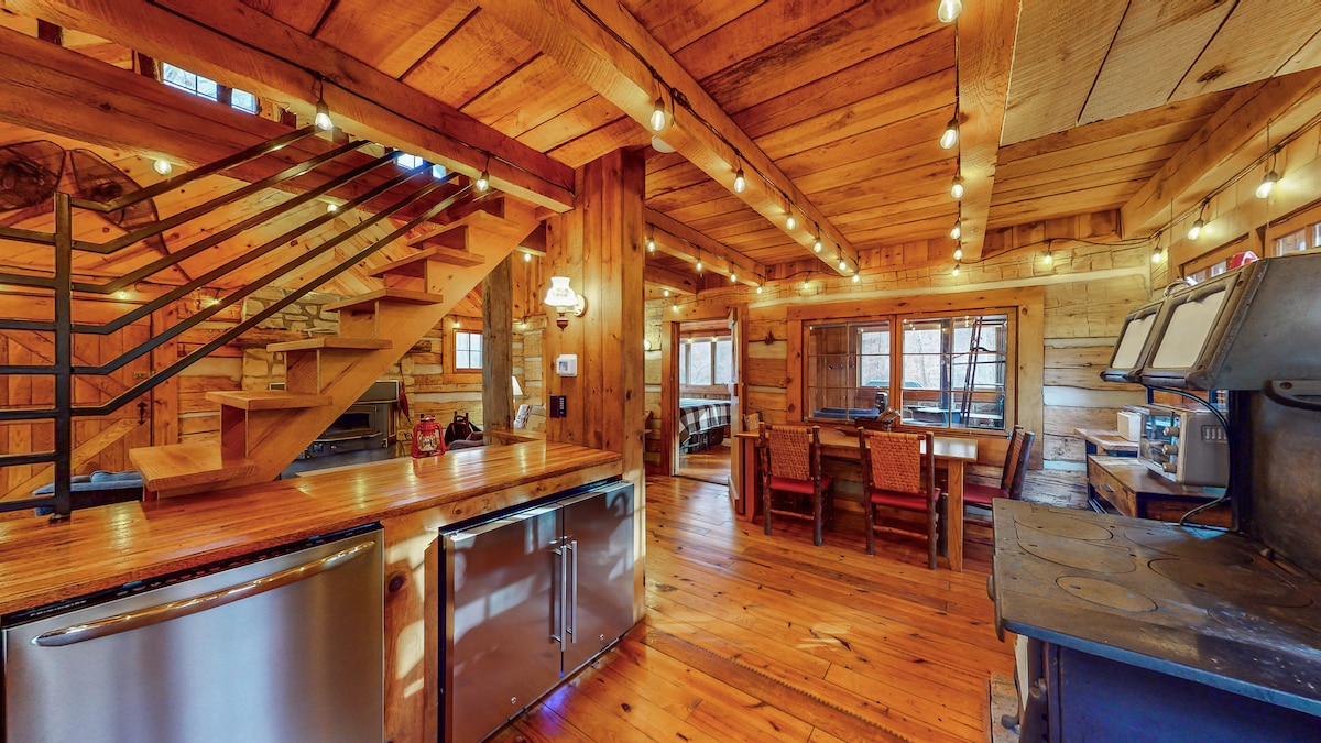 A rustic kitchen and dining area are displayed, featuring wooden beams and a warm wood floor. The space incorporates a spiral staircase, stainless steel appliances, and a large rustic dining table surrounded by several chairs. Ambient lighting is provided by string lights overhead.