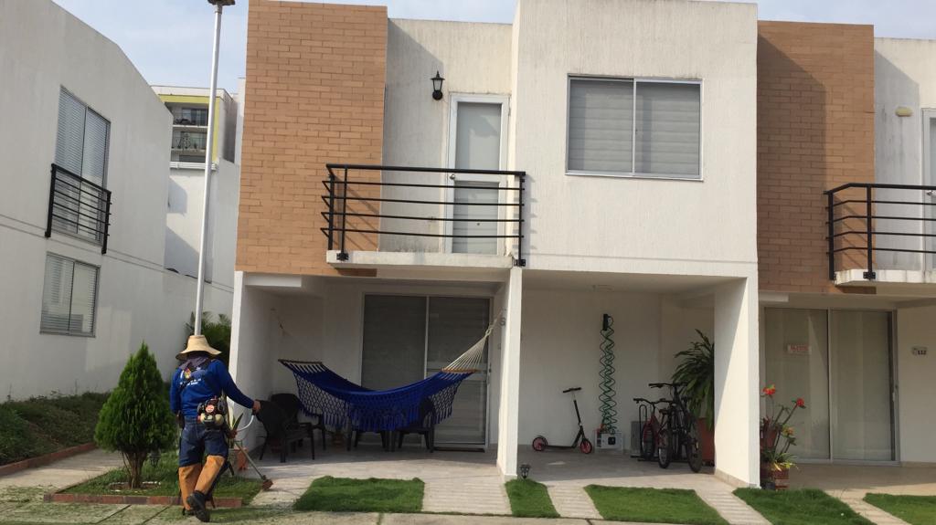 A modern two-story home features a light-colored exterior with contrasting brick accents. A hammock is positioned on the porch, while bicycles are visible nearby. The well-kept garden includes patches of grass and small shrubbery, contributing to a peaceful outdoor space.