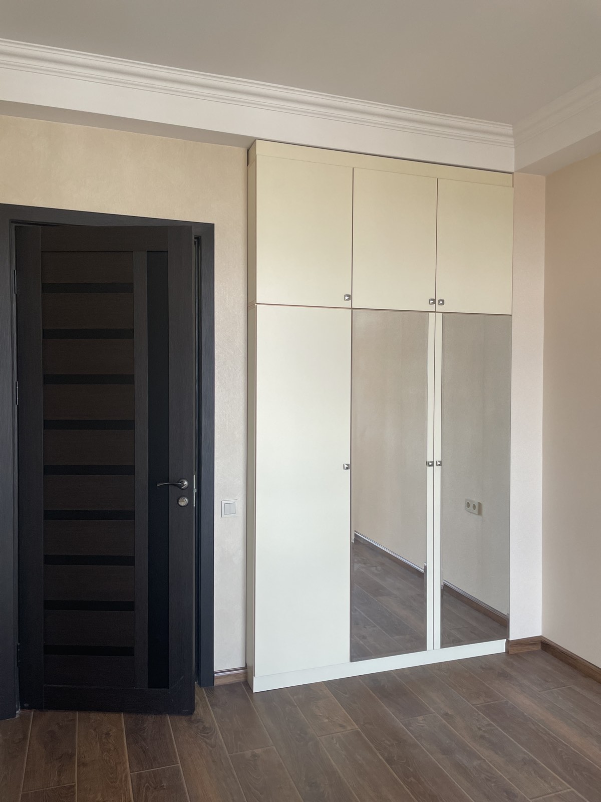 A minimalist interior is depicted, featuring a tall white wardrobe with mirrored doors and multiple storage compartments. A dark wooden door is positioned to the left, contrasting with the light-colored walls and flooring. Natural light illuminates the space, enhancing its clean lines.