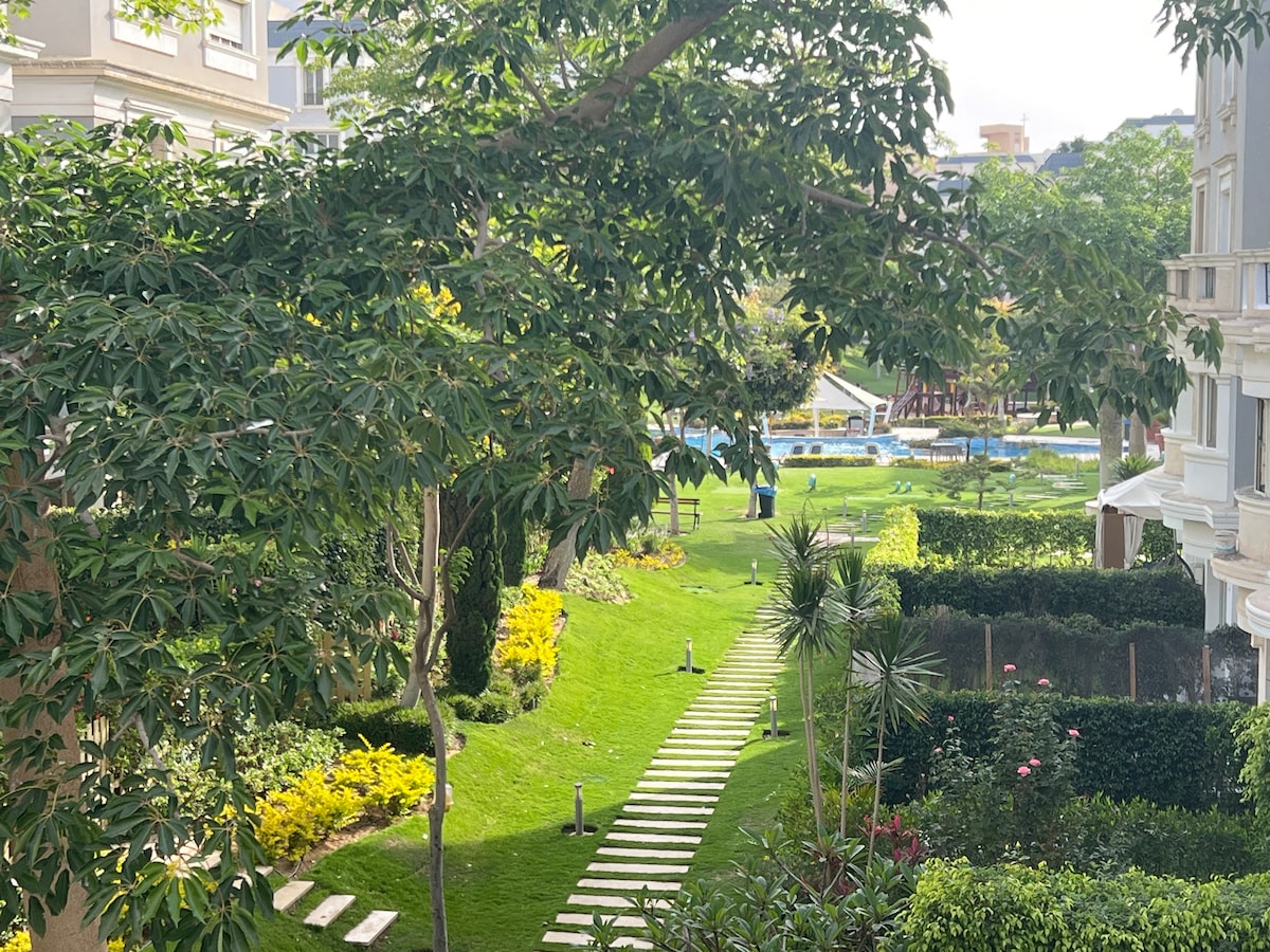 A lush green garden is observed, featuring neatly arranged stepping stones leading through vibrant landscaping. The area is bordered by tall trees and various plants, with a glimpse of a pool area visible in the distance, contributing to a peaceful outdoor environment.