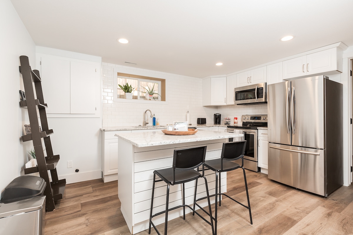 An airy kitchen area is illustrated with white cabinetry and a marble island that includes two black bar stools. Stainless steel appliances, including a refrigerator and microwave, are visible. Natural light enters through a window, enhancing the sleek and functional design.