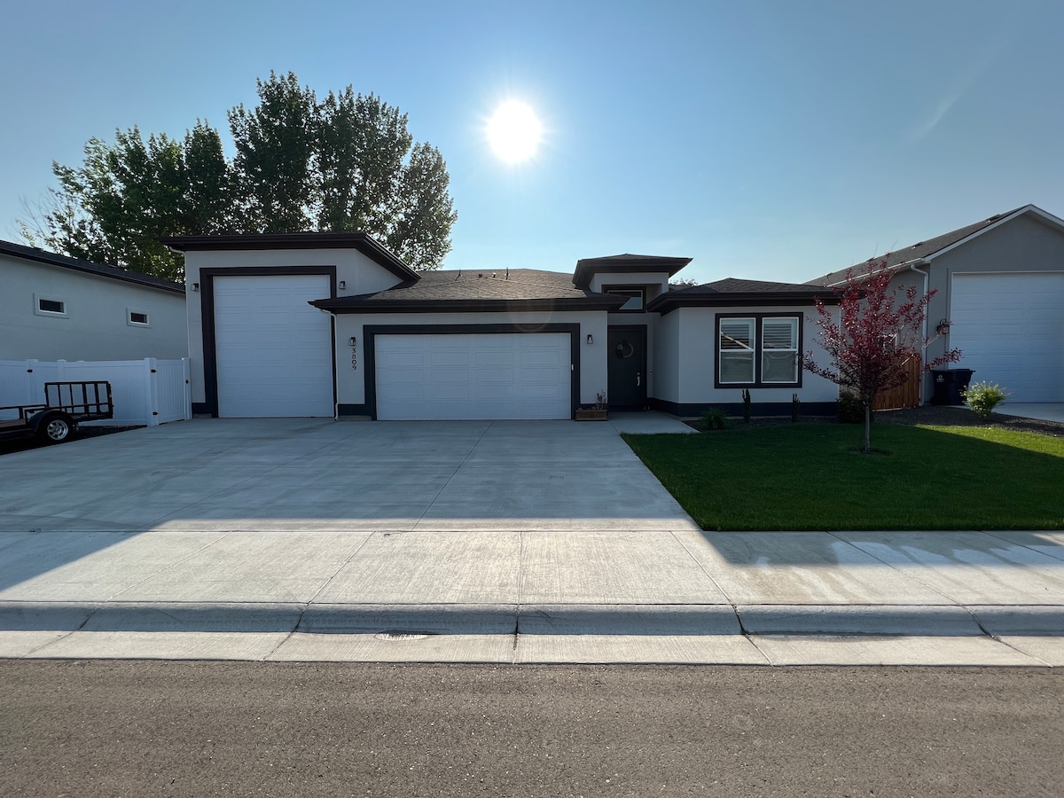 A single-story home is depicted, featuring a contemporary design with a three-car garage. A sunlit front yard, bordered by greenery, presents a clean driveway leading to a well-maintained exterior. The building is accompanied by neighboring residences, contributing to the overall neighborhood appeal.