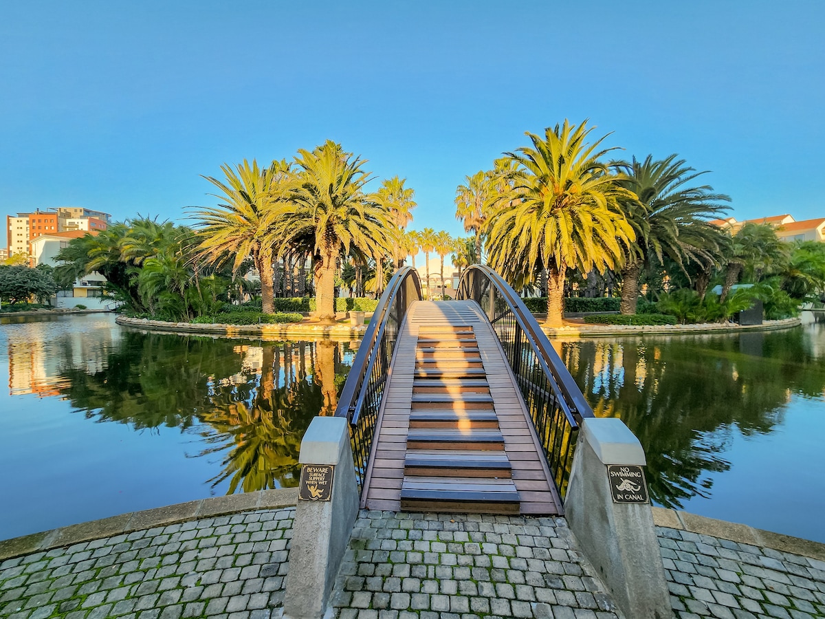 A wooden bridge arches gracefully over still water, leading into a lush, landscaped area filled with palm trees. The reflections of the surrounding greenery and blue sky create a serene atmosphere, inviting guests to explore the tranquil surroundings.