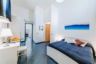 A well-lit bedroom features a spacious bed with decorative pillows against a blue wall art piece. The room includes a flat-screen TV mounted on the wall, a bedside lamp, and a chair. A doorway leads to a separate kitchen area.