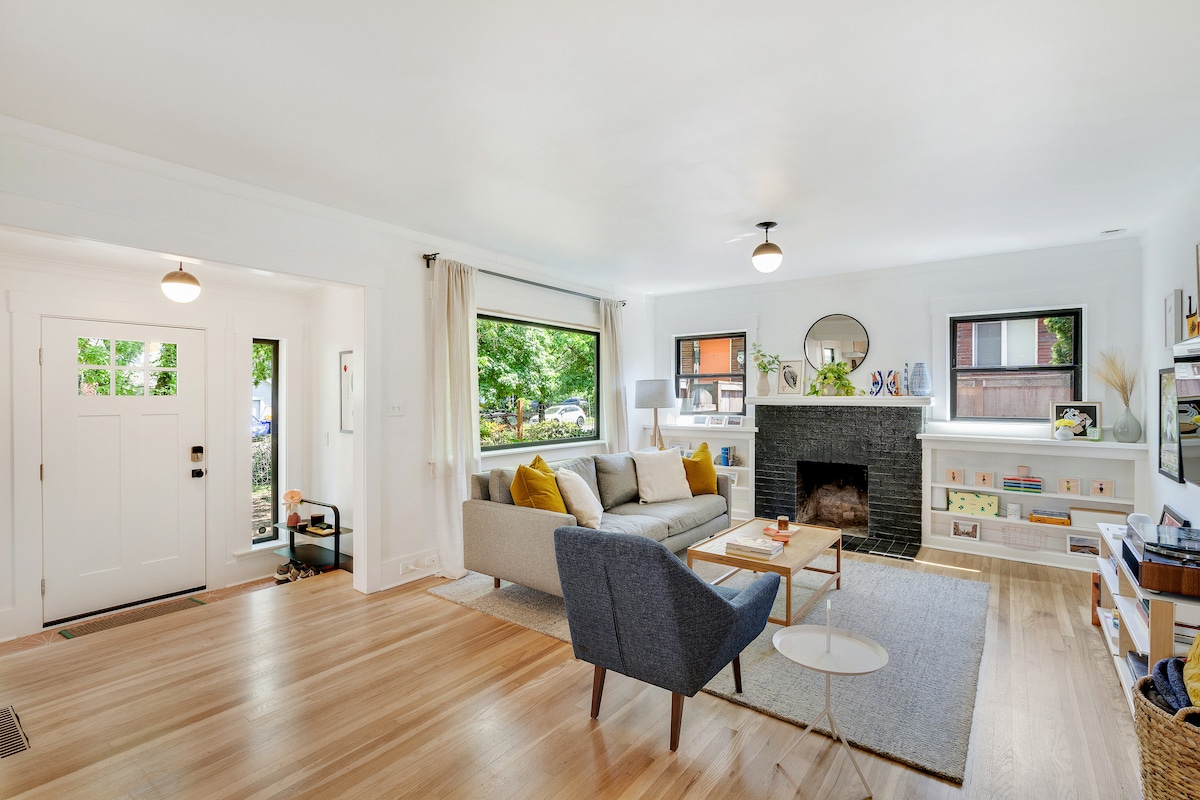 A bright living room featuring light hardwood floors and white walls. A cozy sofa and a stylish armchair are arranged around a coffee table. A fireplace made of black brick stands against one wall, while large windows allow natural light to fill the space.