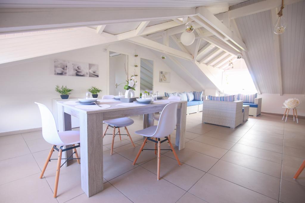 A bright dining area is presented, featuring a spacious table surrounded by white chairs. Natural light floods the room through large windows, highlighting the high ceilings with exposed wooden beams. A comfortable seating arrangement is visible in the background, enhancing the inviting atmosphere.