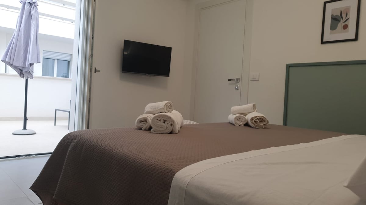 The inviting room features a spacious bed adorned with neatly rolled towels. A wall-mounted television is positioned opposite, with a view of the terrace visible through sliding doors. Natural light enters the room, enhancing the contemporary design highlighted by neutral tones.