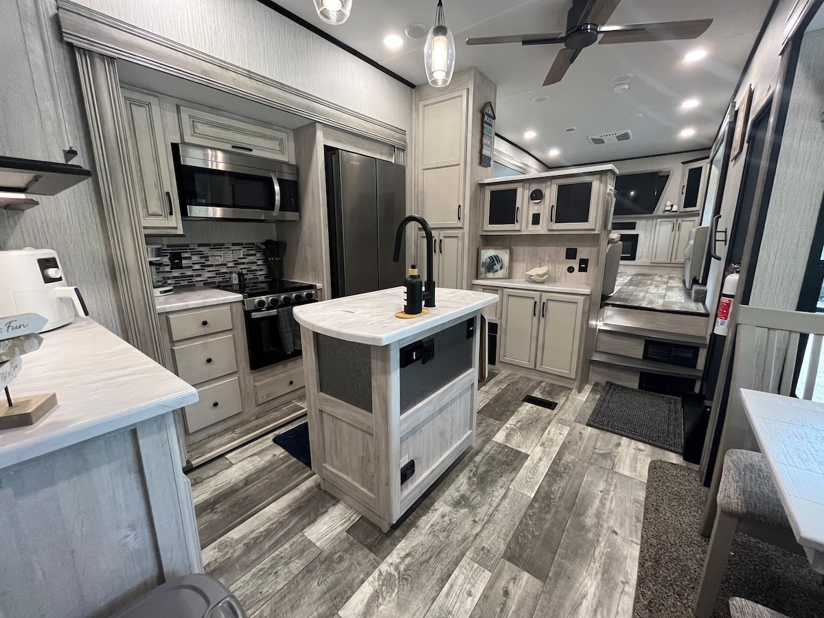 The interior of the camper showcases a modern kitchen area featuring a central island with a double basin sink. Stainless steel appliances, including an oven and refrigerator, are set against light cabinetry. Wood-style flooring extends throughout the space, enhancing the open and airy feel.
