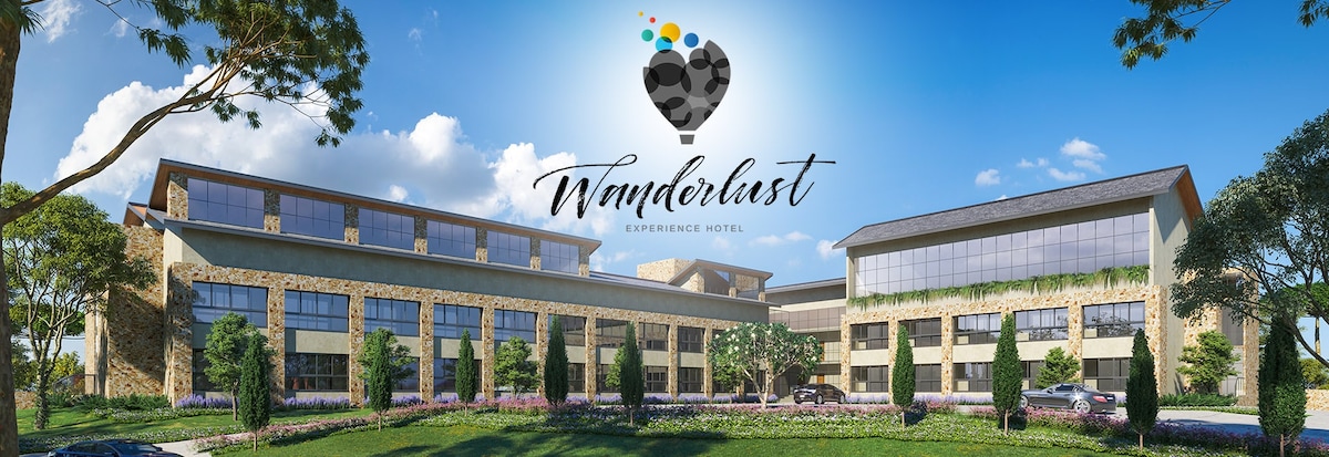 The Wanderlust Experience Hotel is presented against a bright blue sky, showcasing a modern architectural design with stone and wood elements. Lush greenery surrounds the building, complemented by flowering plants in the foreground, creating a welcoming environment.