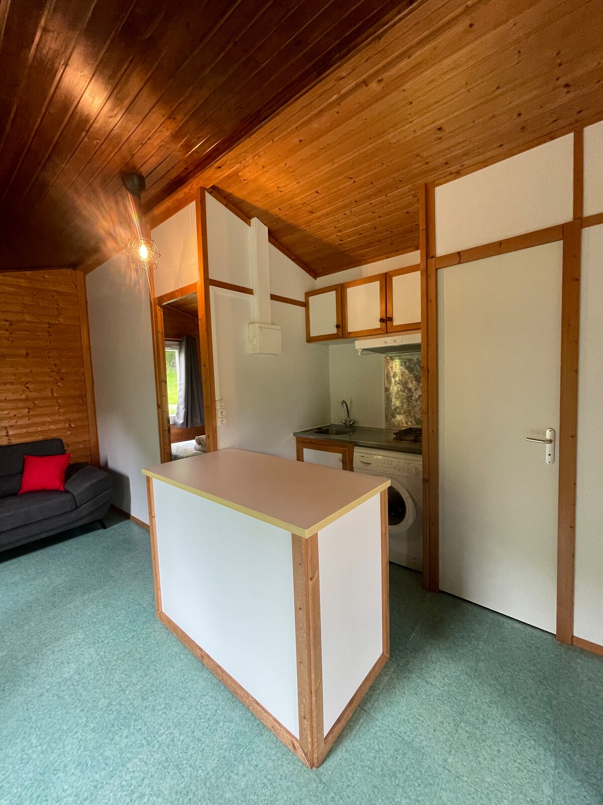 A cozy kitchen area is contained within a wooden interior, featuring a compact cooking space with cabinets and appliances. A neutral-colored counter delineates the kitchen from the living area, where seating is provided by a dark sofa. Natural light enters through nearby windows.