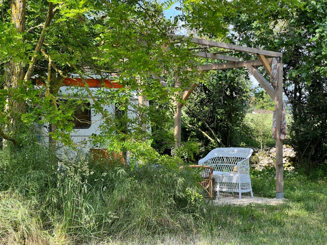 A shaded outdoor seating area features a wooden pergola with a white wicker chair and a small table. Lush greenery surrounds the space, providing a peaceful retreat next to the vintage caravan, which is partially visible in the background.